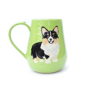 Puede incluir: Taza de cerámica verde claro con una ilustración de un perro corgi negro, blanco y canela. La taza tiene forma redondeada y un asa curva. El perro está sonriendo con la lengua fuera.