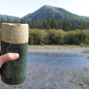 Puede incluir: Una taza de cer&aacute;mica con la parte superior beige moteada y la parte inferior verde oscuro. La taza est&aacute; siendo sostenida por una mano frente a un fondo borroso de un r&iacute;o y &aacute;rboles.