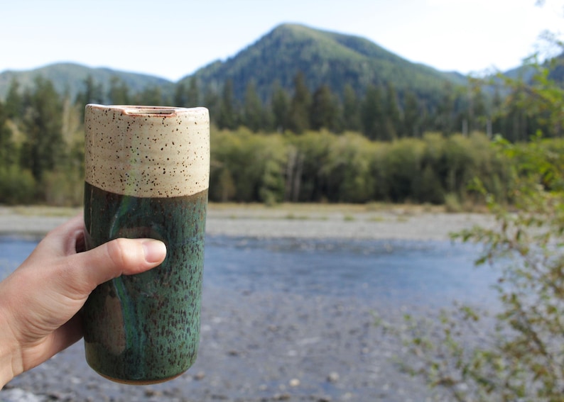 Puede incluir: Una taza de cer&aacute;mica con la parte superior beige moteada y la parte inferior verde oscuro. La taza est&aacute; siendo sostenida por una mano frente a un fondo borroso de un r&iacute;o y &aacute;rboles.