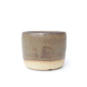 Puede incluir: Una taza cilíndrica de cerámica con un esmalte de dos tonos. La parte superior tiene un acabado liso de color marrón oliva, mientras que la sección inferior es de color crema blanquecino con motas. El recipiente tiene un borde recto.