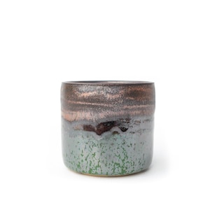 May include: A cylindrical ceramic cup featuring a two-tone glaze. The upper portion displays a dark, metallic bronze and brown finish, while the lower section transitions into a mottled, textured green and grey glaze with a speckled appearance.