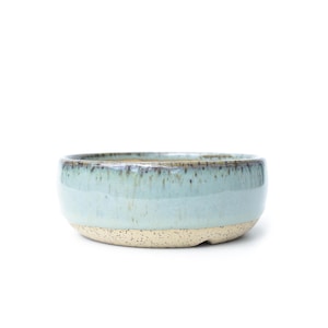 May include: A handmade ceramic bowl with a light blue glaze and a speckled, beige base. The bowl has a rounded shape and a smooth, glossy finish. The rim of the bowl has a darker, textured edge. This bowl is perfect for serving food or as a decorative piece.