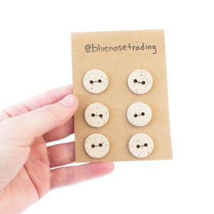 Könnte beinhalten: Sechs gesprenkelte, cremefarbene Keramikknöpfe mit jeweils zwei Löchern werden auf einer braunen Karte präsentiert. Auf der Karte steht der Text "@bluenosetrading". Die Knöpfe sind rund und haben ein gesprenkeltes Design.