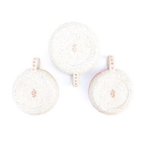 May include: Three speckled ceramic mugs, viewed from the bottom, arranged against a white background. The mugs have a light pink rim and handle, with a speckled off-white exterior. Each handle is adorned with small pink dots.
