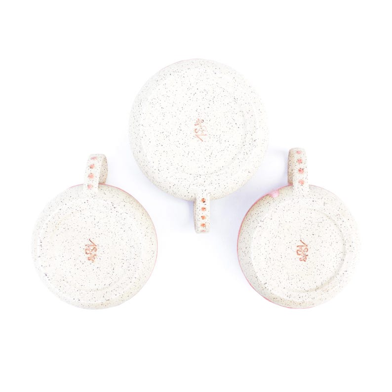 May include: Three speckled ceramic mugs, viewed from the bottom, arranged against a white background. The mugs have a light pink rim and handle, with a speckled off-white exterior. Each handle is adorned with small pink dots.