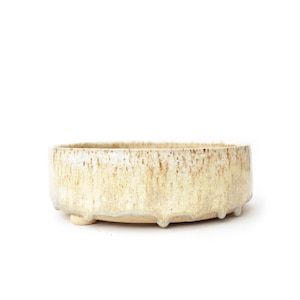 May include: A round ceramic bowl with a low profile and three small feet. The exterior features a textured, speckled cream and beige glaze that drips down the sides, creating a layered, variegated appearance against the base clay colour.