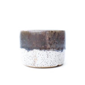 May include: A ceramic, cylindrical planter with a speckled white base and a dark brown, glazed top. The planter has a smooth, rounded shape and is set against a white background. The planter is suitable for small plants.