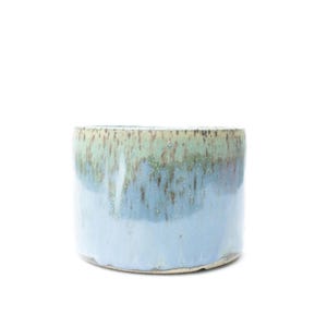 May include: A cylindrical ceramic planter featuring a reactive glaze finish. The upper portion displays a mottled sage green and brown tone, which transitions into a smooth, solid light blue color on the lower half of the vessel's exterior.