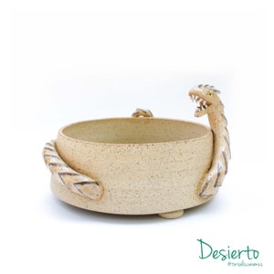 以下が含まれることがあります： ベージュのセラミックボウルで、2つのドラゴン型のハンドルが付いています。ボウルは斑点模様のテクスチャで、丸い底をしています。ドラゴンのハンドルは鱗で装飾されており、薄い茶色の釉薬がかけられています。