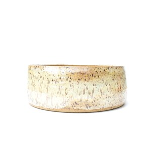 May include: A handcrafted ceramic bowl with a speckled, glazed finish. The bowl features a wide, shallow design with a neutral color palette of cream, tan, and brown. The exterior has a textured appearance, adding to its rustic charm.