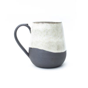 May include: A handmade ceramic mug with a two-tone design. The bottom half is a dark charcoal gray, while the top half is a speckled off-white. The mug has a curved handle and a smooth, rounded shape, perfect for coffee or tea.