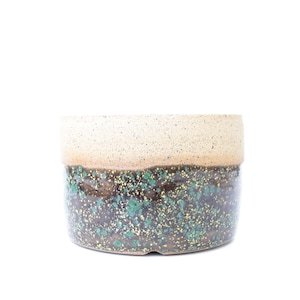 May include: A cylindrical ceramic planter featuring a two-tone glaze. The upper portion is a speckled cream color, while the lower section displays a dark brown base with vibrant green and gold flecks throughout the textured surface.