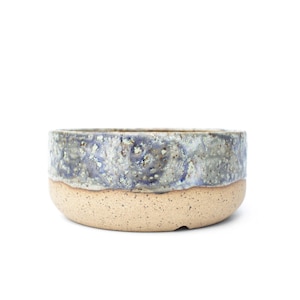 May include: A handcrafted ceramic bowl with a speckled, light brown base and a textured, blue-grey rim. The bowl has a rounded shape and is suitable for serving food or as a decorative piece. The bowl's dimensions are not visible.