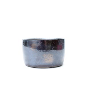 May include: A cylindrical ceramic bowl with a dark, glossy finish featuring subtle metallic bronze speckles throughout the surface. The bowl has a smooth, rounded rim and a flat base, presenting a deep, reflective appearance with a dark color palette.