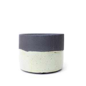 Puede incluir: Una maceta cilíndrica de cerámica con un diseño de dos tonos, que presenta una sección superior en negro carbón mate y una sección inferior en blanco roto con motas. El recipiente tiene una base plana y paredes verticales rectas, mostrando un contraste marcado entre la parte superior oscura y texturizada y el acabado más claro y ligeramente moteado de la base.