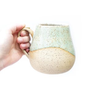 Peut inclure: Une tasse en céramique de couleur vert clair et beige avec un glaçage moucheté. La tasse a un corps arrondi et une anse.