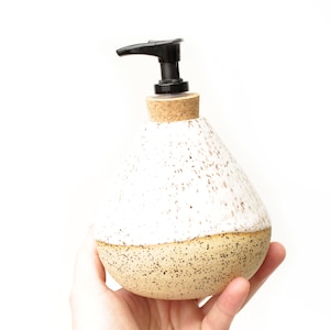 May include: A ceramic soap dispenser with a teardrop shape, featuring a white speckled upper section and a tan speckled lower section. The top includes a cork collar and a black plastic pump mechanism for dispensing liquid soap or lotion.