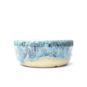 May include: A ceramic bowl featuring a speckled cream-colored base and a textured, drippy glaze in shades of turquoise and dark blue that cascades down from the rim, creating a distinct contrast between the smooth upper section and the base.