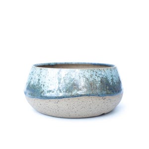 May include: A ceramic bowl with a speckled, light beige base and a top section with a blue and green glaze. The bowl has a rounded shape and a wide opening, suitable for various uses.