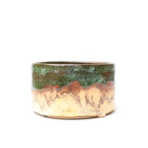 May include: A cylindrical ceramic bowl featuring a two-tone glaze. The upper portion is a deep forest green with dark speckles, while the lower section transitions into a textured, creamy beige base with a visible drip pattern along the rim.