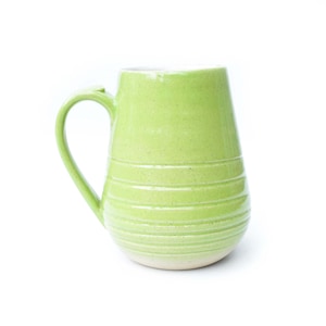 May include: A handmade, light green ceramic mug with a large handle. The mug has horizontal, textured lines around the body. The bottom of the mug is a lighter shade of green. The mug is set against a white background.