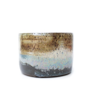 Puede incluir: Una taza cilíndrica de cerámica con un esmalte de varios tonos. La sección superior muestra un acabado moteado en marrón y bronceado, que transiciona a una banda iridiscente de color azul grisáceo y termina en una base gris oscuro.