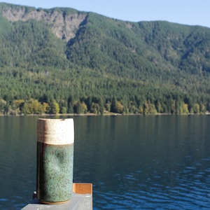 Puede incluir: Una taza de cer&aacute;mica con la parte superior de color marr&oacute;n claro y la parte inferior de color verde oscuro se encuentra sobre una superficie de madera frente a un lago y monta&ntilde;as.