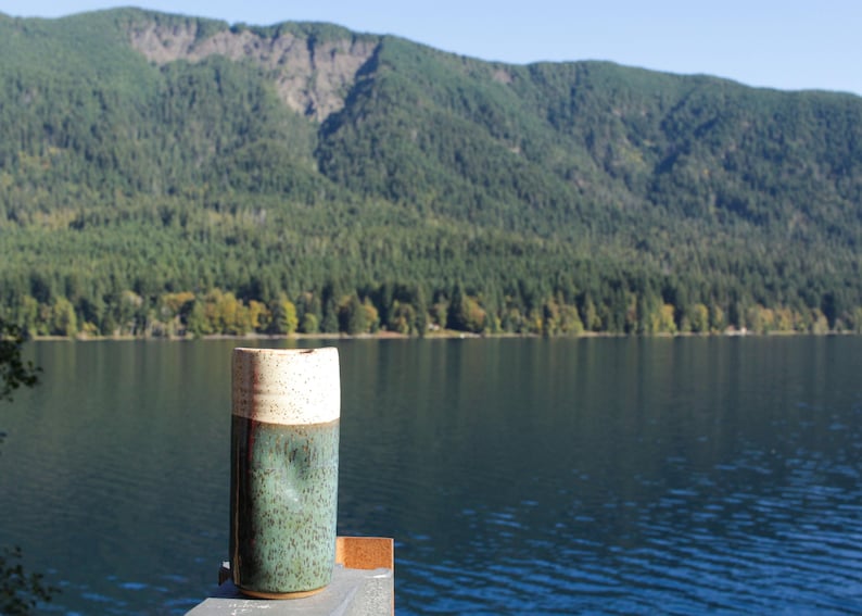 Puede incluir: Una taza de cer&aacute;mica con la parte superior de color marr&oacute;n claro y la parte inferior de color verde oscuro se encuentra sobre una superficie de madera frente a un lago y monta&ntilde;as.