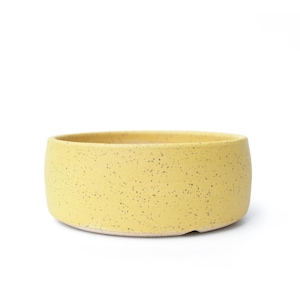 May include: A round, yellow ceramic bowl with a speckled texture. The bowl has a low profile and a wide opening, suitable for serving or display. The exterior is a solid, muted yellow with small, dark speckles.