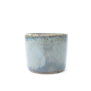 May include: A small, cylindrical ceramic planter with a light blue and grey glaze. The handmade pot has a slightly textured surface and a dark rim. Suitable for small plants and succulents.