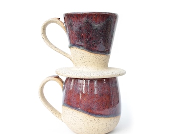 May include: A ceramic pour-over coffee dripper stacked on top of a matching ceramic mug. Both pieces feature a speckled cream-colored base and a deep, glossy burgundy glaze on the upper sections. Each item includes a curved handle for easy gripping.