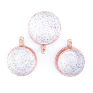 May include: Overhead shot of three handmade ceramic mugs. The mugs have a speckled white interior and a pinkish-red rim and handle. The handles are small and rounded. The mugs are arranged on a white background.