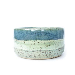 May include: A ceramic bowl with a speckled, layered design. The bowl features bands of blue, green, and off-white colors. The bowl has a rounded shape and a glossy finish, suitable for home decor or serving.