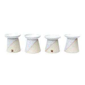 May include: Four ceramic pet bowls. Each bowl has a speckled white top and a beige base with a diagonal color split. A small paw print is on the base of each bowl. The bowls are elevated.