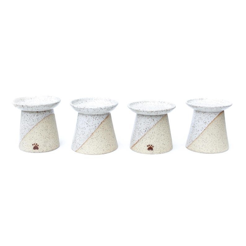 May include: Four ceramic pet bowls. Each bowl has a speckled white top and a beige base with a diagonal color split. A small paw print is on the base of each bowl. The bowls are elevated.