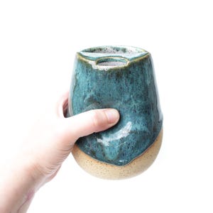 May include: A ceramic vessel with a rounded base and a tapered top featuring a central opening. The upper portion is finished in a glossy, speckled teal glaze, while the lower section displays a matte, speckled beige clay texture.