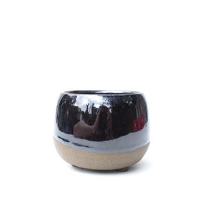 May include: A small, rounded ceramic planter featuring a glossy, dark black glazed upper section and a contrasting, unglazed matte tan base. The vessel has a wide, open top and a smooth, curved silhouette, resting on a small, integrated footed base.