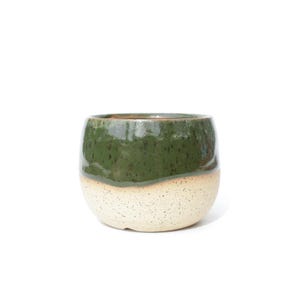 May include: A rounded ceramic planter featuring a two-tone design with a glossy dark forest green glaze on the upper portion and a speckled cream-colored base. The vessel has a wide, open top and a smooth, curved silhouette without handles.