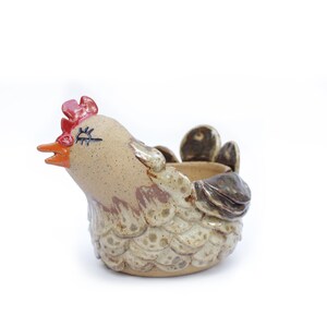 May include: A ceramic chicken planter with a brown and white speckled glaze. The chicken has a red comb and wattle, and a closed eye. The planter has a small opening on the back for drainage.