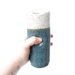 May include: A ceramic vase with a speckled white top and a speckled teal bottom. The vase is held in a hand.