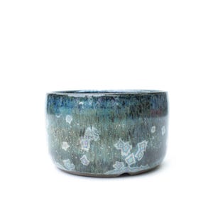 May include: A cylindrical ceramic bowl featuring a deep blue and teal crystalline glaze. The surface displays a variegated texture with distinct, star-like white and light blue crystal formations scattered across the dark, glossy exterior finish.