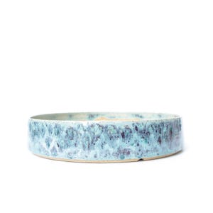 May include: A low, round ceramic bowl with a textured, mottled blue and teal glaze. The bowl has a light beige interior and a simple, minimalist design, perfect for serving or display.