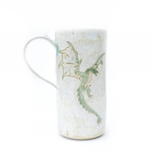 May include: A tall, off-white ceramic mug with a curved handle. The mug features a hand-painted green dragon design on the front. The mug has a textured surface and a light blue accent on the handle.
