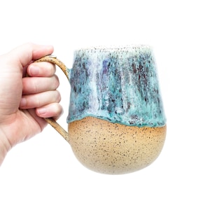 Peut inclure: Une tasse en céramique avec une base arrondie, présentant une finition beige tachetée sur la moitié inférieure et un vernis brillant bleu sarcelle et bleu foncé coulant sur la moitié supérieure. La tasse possède une anse incurvée sur le côté.
