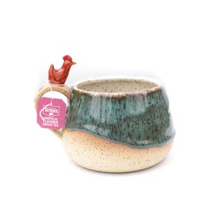 May include: A ceramic teacup with a green and brown glaze and a small red chicken figurine on the handle. The teacup has a speckled finish and a tag attached to the handle that reads "T.G.I. Benner Tropical Flavored Green Tea".