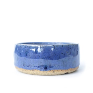 Puede incluir: Un cuenco de cerámica hecho a mano con forma redondeada. El cuenco presenta un esmalte azul brillante y moteado en el exterior y una base beige natural sin esmaltar. Adecuado para servir alimentos o como pieza decorativa.