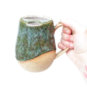 May include: A ceramic mug featuring a two-tone glaze with a glossy dark green top and a speckled beige bottom. The mug has a curved handle and a unique integrated notch at the rim, designed for resting a tea bag string or similar small item.