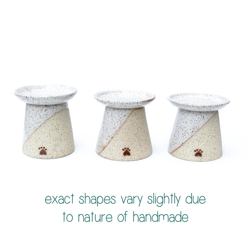 May include: Three ceramic pet bowls with a speckled white top and a beige and white diagonal design on the base. Each bowl has a paw print and the text "exact shapes vary slightly due to nature of handmade".