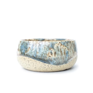 May include: A ceramic bowl with a speckled, off-white base and a top section with a blue, brown, and beige glaze. The bowl has a rounded shape and a smooth, glossy finish. The bowl is suitable for serving food.