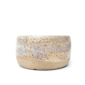 May include: A cylindrical ceramic bowl featuring a two-tone design. The upper portion displays a mottled glaze with streaks of cream, beige, and soft brown. The lower section is composed of unglazed, speckled tan clay with a natural, matte texture.